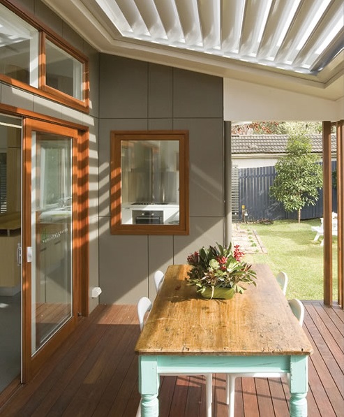 Close view of timber deck furnished with rustic table and contemporary chairs leads through large glass sliding doors to living room and kitchen. Deck is shaded by adjustable louvres. There is a grassed yard beyond the deck.
