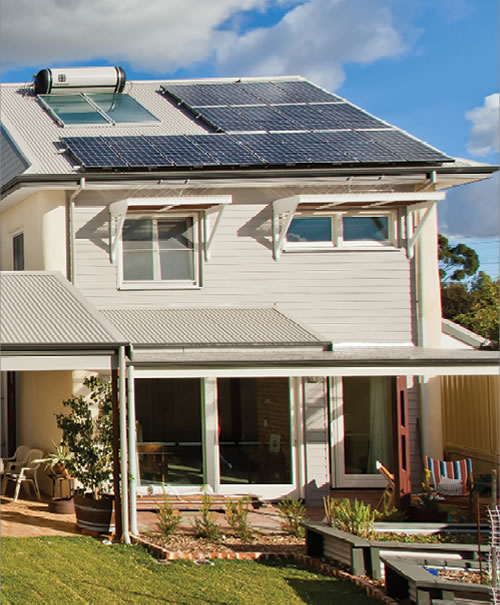 A view of a two story house from the backyard showing vegetable beds, mulched garden, a pergola in front of large glass doors to the living room, fixed awnings over upstairs windows and photovoltaic solar panels and solar hot water service on the roof.