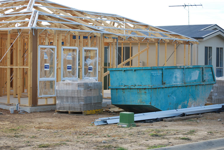 A building site with a house in its early stages of construction. There is a concrete slab, timber faming and some front windows installed. In the foreground a there is a large skip for waste materials. 