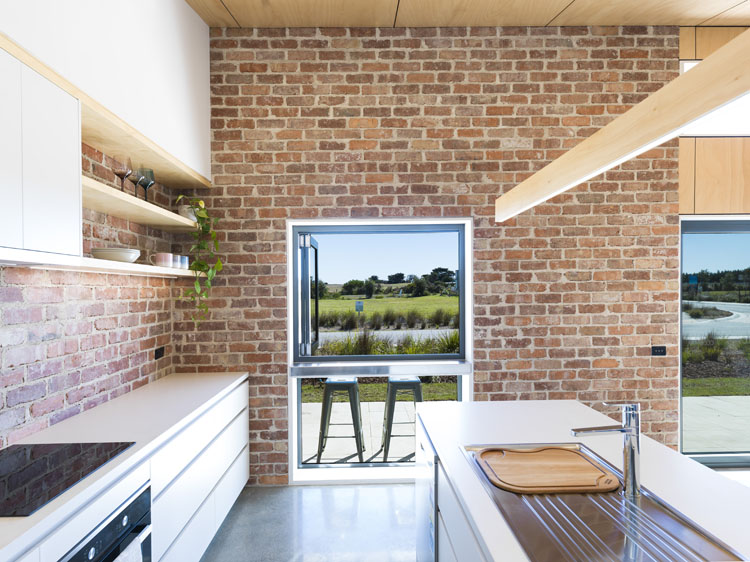A photo shows an exposed brick wall at the end of a modern galley kitchen, with a large, bi-fold window with a view across a country landscape.  