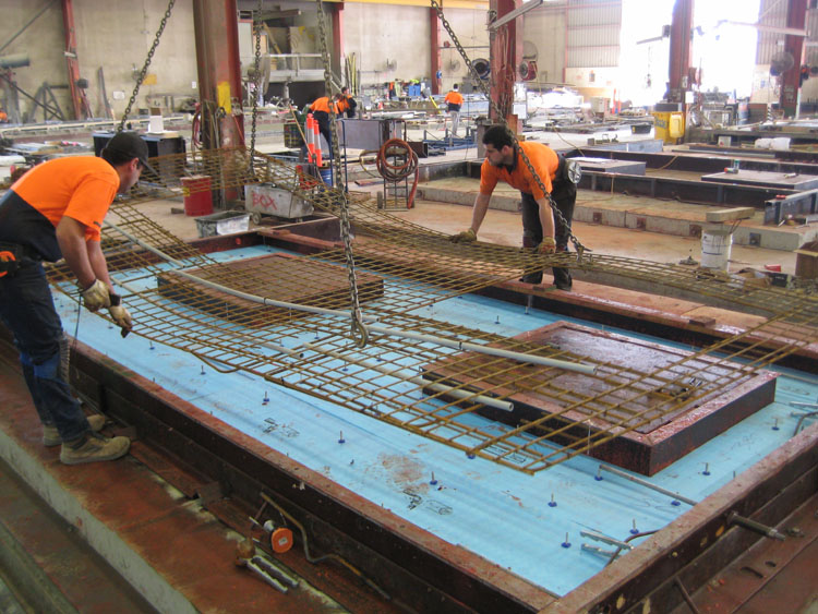 Photo of the construction of a precast concrete panel in the factory. The panels have the steel reinforcement frame, and cut-outs for windows and services. The panels are ready to be poured.